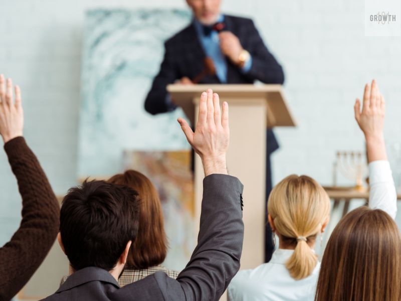 A group of people raising their hands in a bidding scenario, symbolizing the auction process, with a speaker conducting the auction from a podium.