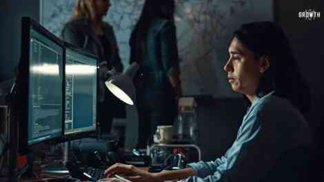 A side-profile candid shot in a dimly lit startup office after hours. A delivery lead sits at a cluttered desk with two glowing monitors reflecting on their face, showing deep focus and slight fatigue. In the background, out of focus, two engineers are seen in a heated but quiet discussion near a whiteboard filled with messy diagrams. The lighting is natural and moody, coming from the desk lamps and screen glow, creating a raw, unpolished documentary feel. There are half-empty coffee mugs and tangled cables on the desk, capturing the high-stakes pressure of a scaling company.
