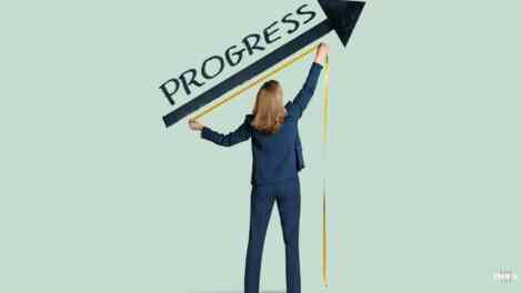 Image of a woman using a measuring tape on an arrow that is underneath the word progress written on the wall.