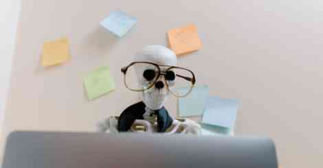 Skeleton wearing glasses and a tie working at a desk with sticky notes on the wall, symbolizing burnout from a 9-to-5 office job.